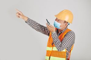 Contractor in medical mask using walkie-talkie when controlling work on construction site